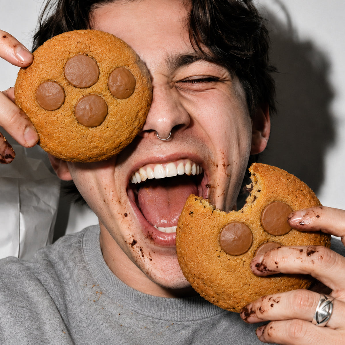 Lachender junger Mann mit dunklen Haaren und Septumring hält zwei große THC Schokoladen-Cookies vor sein Gesicht, einen davon wie eine Augenklappe. Auf Gesicht und Händen sind Schokoladenkrümel zu sehen.