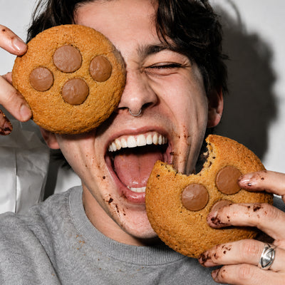 Lachender junger Mann mit dunklen Haaren und Septumring hält zwei große THC Schokoladen-Cookies vor sein Gesicht, einen davon wie eine Augenklappe. Auf Gesicht und Händen sind Schokoladenkrümel zu sehen.